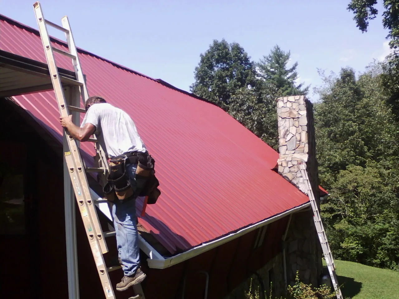 Roofer on a ladder beside a red metal roof and stone chimney — Soffit & Fascia Repair by Roofing Brothers in Arena